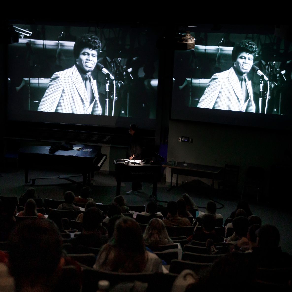 Students watch an archival video clip on the screen of a darkened lecture hall