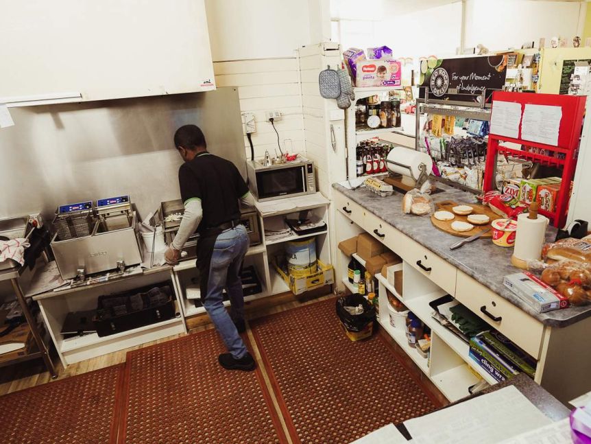 A man in jeans and a black apron stands behind a grill and fryer in a small kitchen.