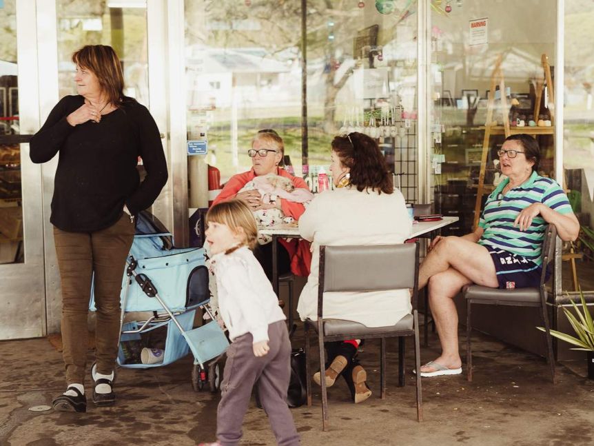 A woman stands next to a table of women chatting sitting down outside a glass shopfront.