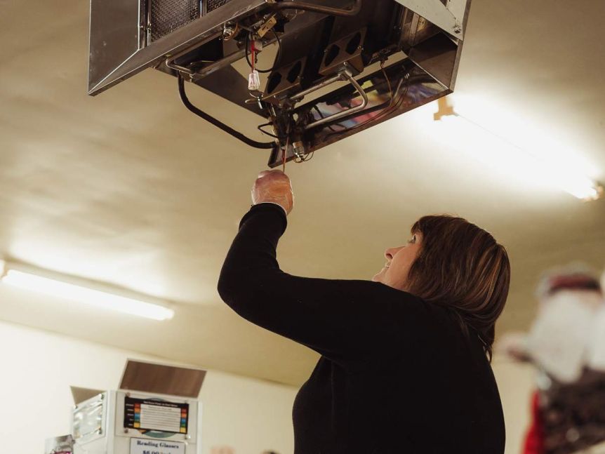 A woman withbrown hair dressed in a black shirt stands on a chair adjusting a ceiling exhaust inside.