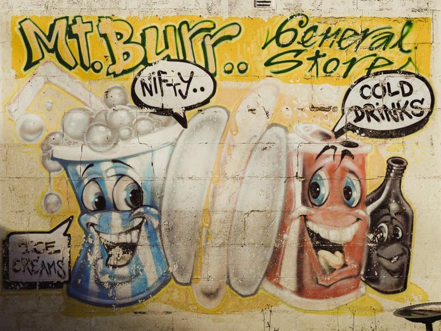 A mural with the words 'Mt Burr General Store' painted in green and yellow with two cartoon smiling soda cans below it. 