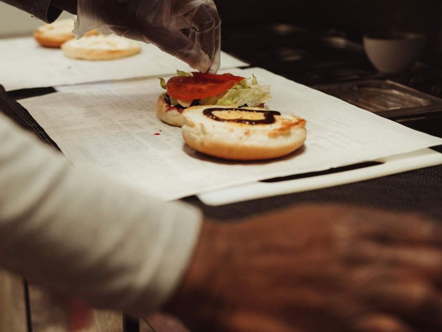 A hand in a plastic glove places a slice of tomato on an open burger bun with sauce and lettuce already on it.